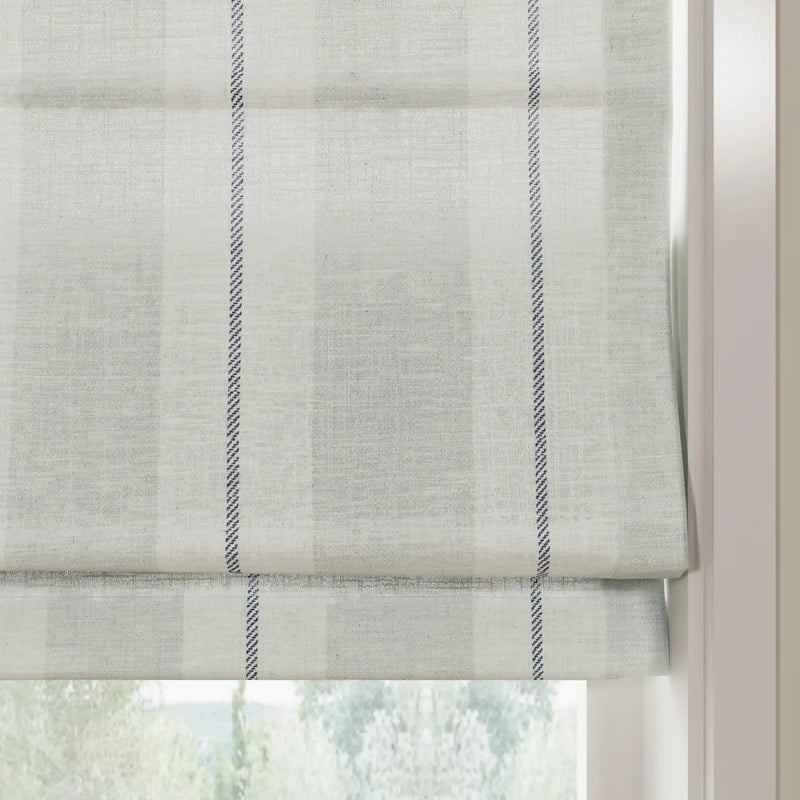 A light grey linen-blend Roman blind with a subtle plaid texture and prominent vertical dark blue dashed stripes, partially lowered in a window revealing a blurred green outdoor view