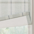 Textile Roman window blind featuring a light gray subtle grid pattern and bold vertical black and white zigzag stripes partially unfurled against a blurred green foliage background