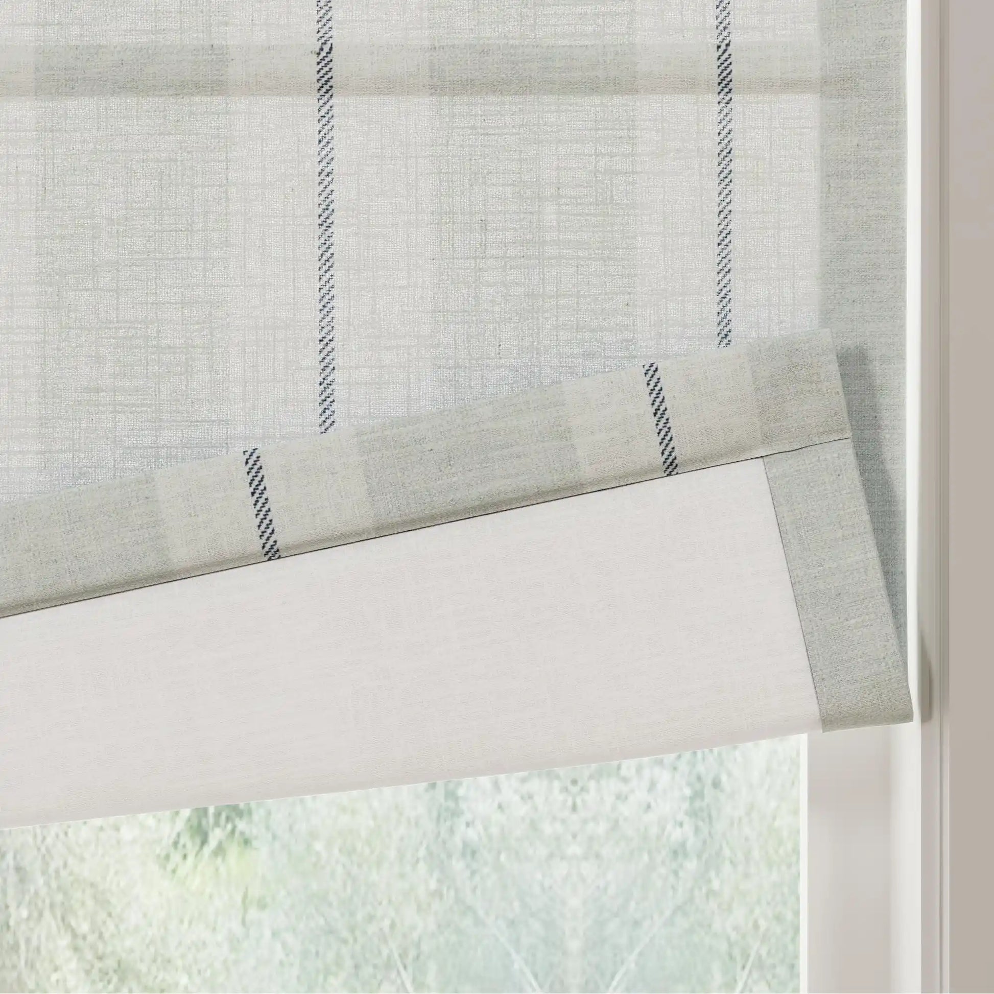 Textile Roman window blind featuring a light gray subtle grid pattern and bold vertical black and white zigzag stripes partially unfurled against a blurred green foliage background