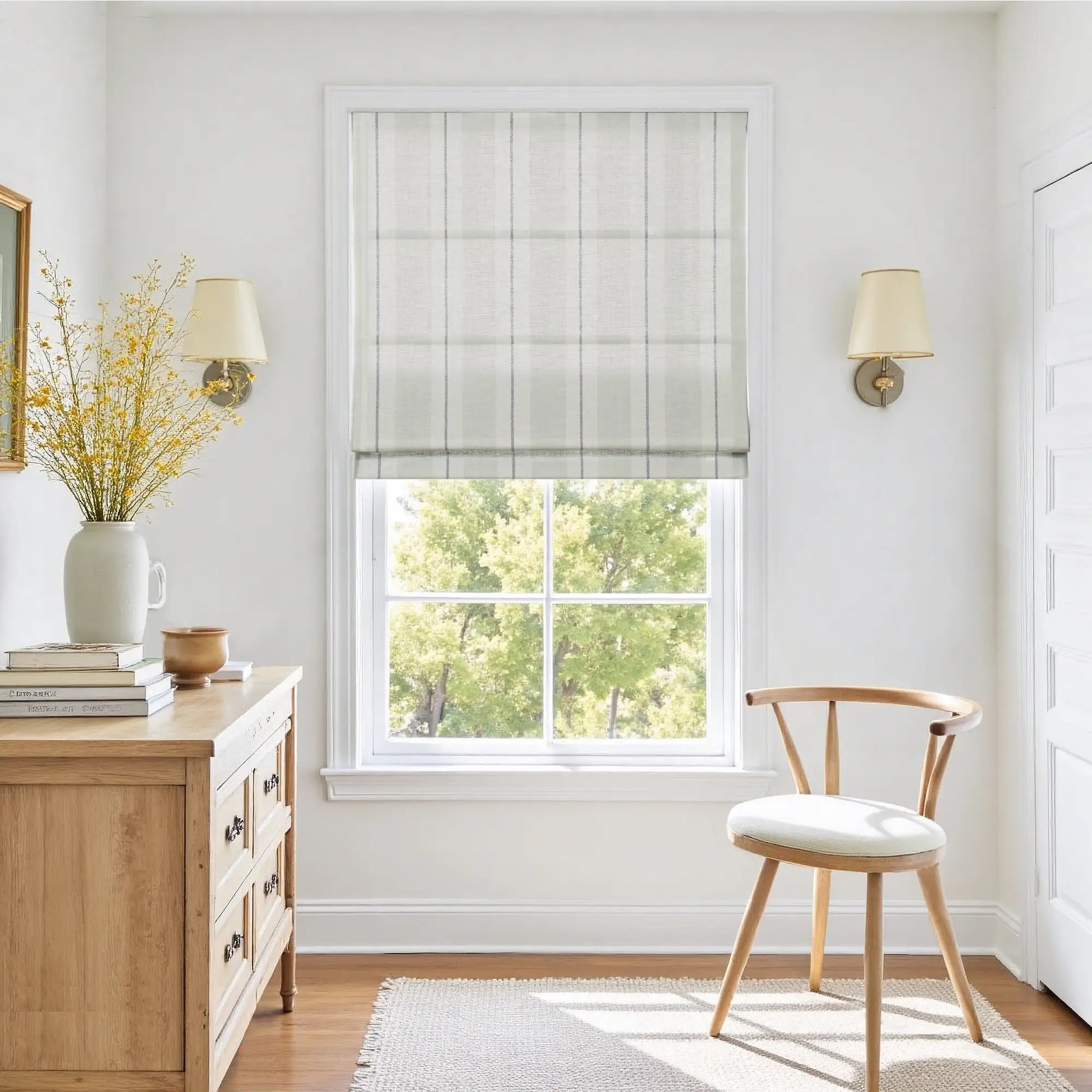 A bright room interior with a window featuring a striped roman shade, a light wood dresser holding a vase of vibrant yellow flowers and stacked books, and a natural wood chair on a textured cream rug