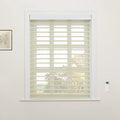 Interior photograph of a window showcasing creamy beige zebra roller Blinds, a white remote control on the adjacent wall, and a framed botanical print to the left, with blurred autumn trees visible beyond