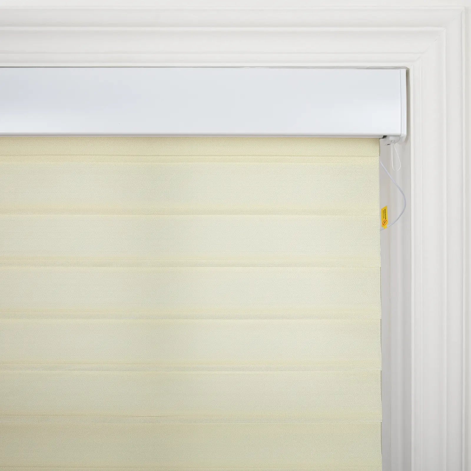 Cream zebra window Blinds with alternating sheer and opaque fabric stripes and a white control cord with a small yellow safety tag set in an ornate white window frame