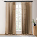Beige linen-look pinch pleat drapes on a black rod framing a white-paned window with blurred greenery, beside a ribbed wooden side table featuring a black vase and gold candle holder