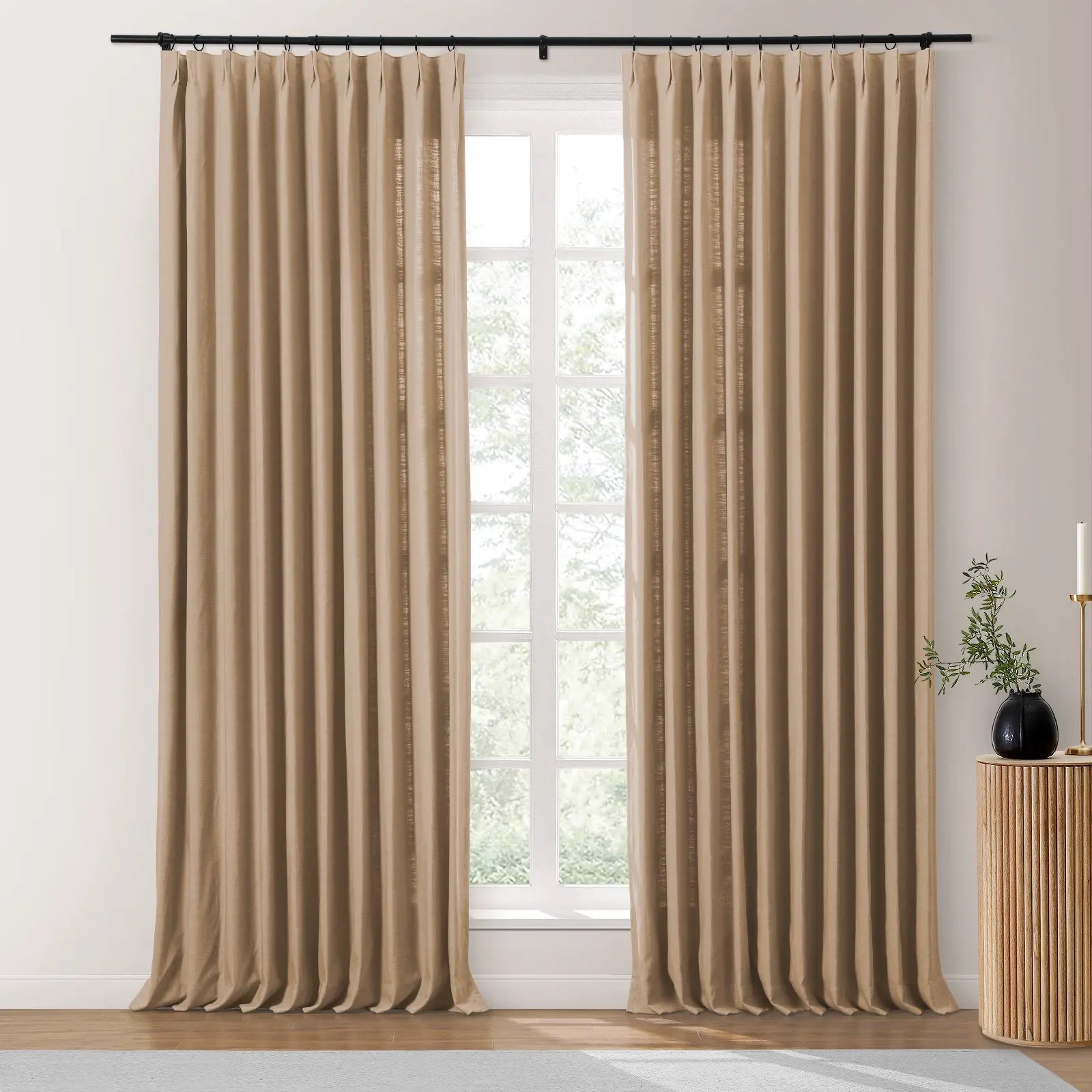 Beige linen-look pinch pleat drapes on a black rod framing a white-paned window with blurred greenery, beside a ribbed wooden side table featuring a black vase and gold candle holder