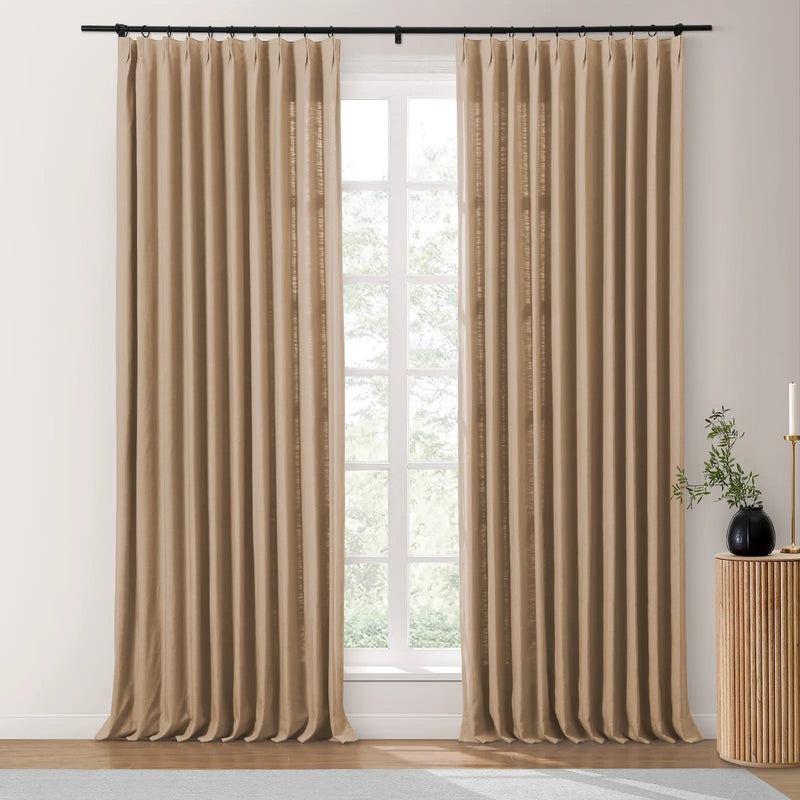 Beige linen-look pinch pleat drapes on a black rod framing a white-paned window with blurred greenery, beside a ribbed wooden side table featuring a black vase and gold candle holder