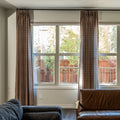 Digital photograph of a living room interior with large windows showcasing brown patterned drapes a dark brown leather armchair and a vibrant backyard view of a wooden fence and green trees