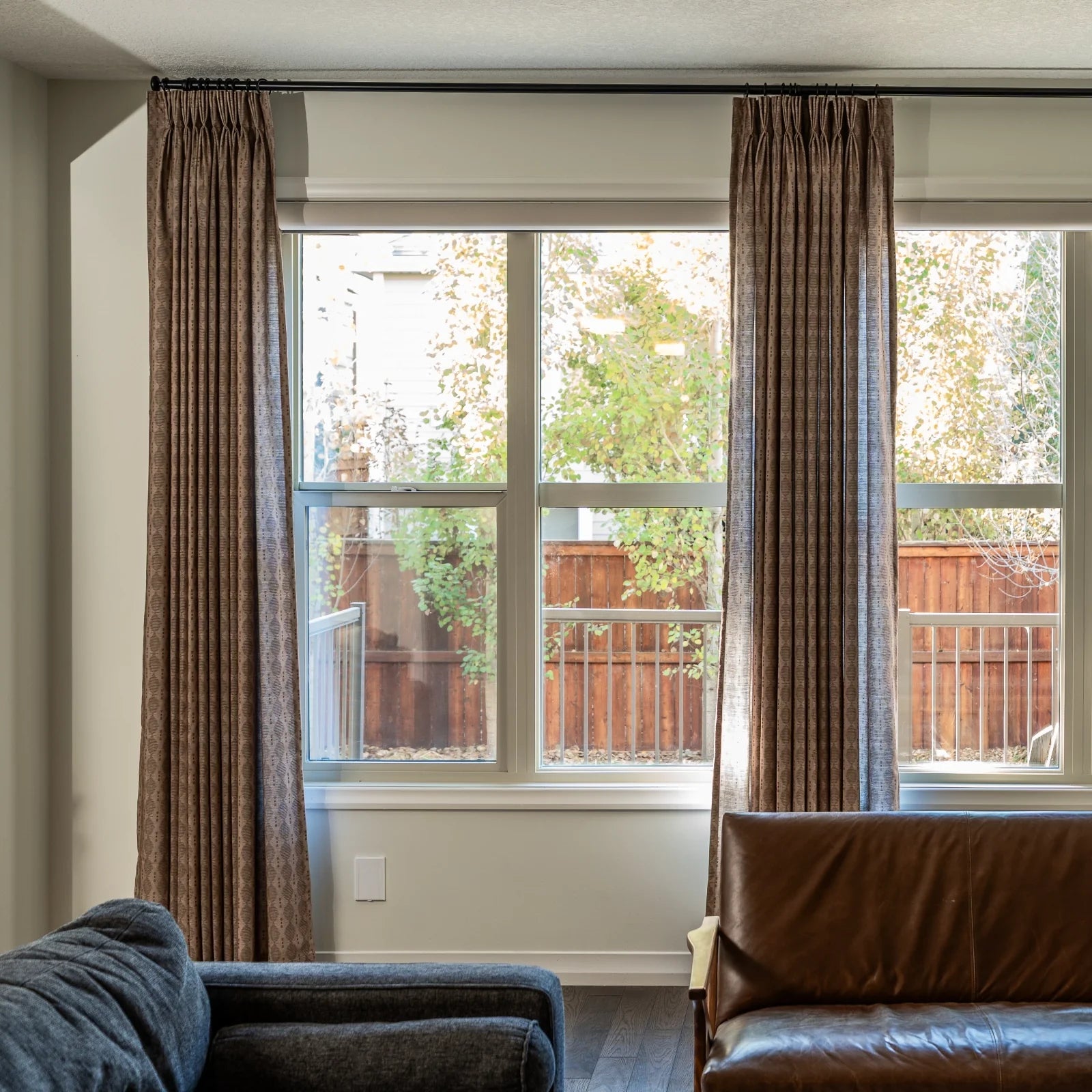 Digital photograph of a living room interior with large windows showcasing brown patterned drapes a dark brown leather armchair and a vibrant backyard view of a wooden fence and green trees