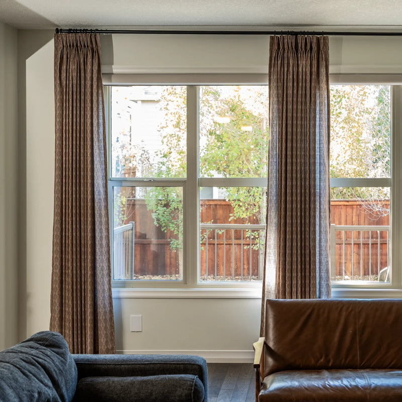 Digital photograph of a living room interior with large windows showcasing brown patterned drapes a dark brown leather armchair and a vibrant backyard view of a wooden fence and green trees