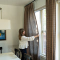 A woman in a white textured blouse adjusts patterned brown curtains in a brightly lit dining room with a vibrant yellow boat painting on the wall