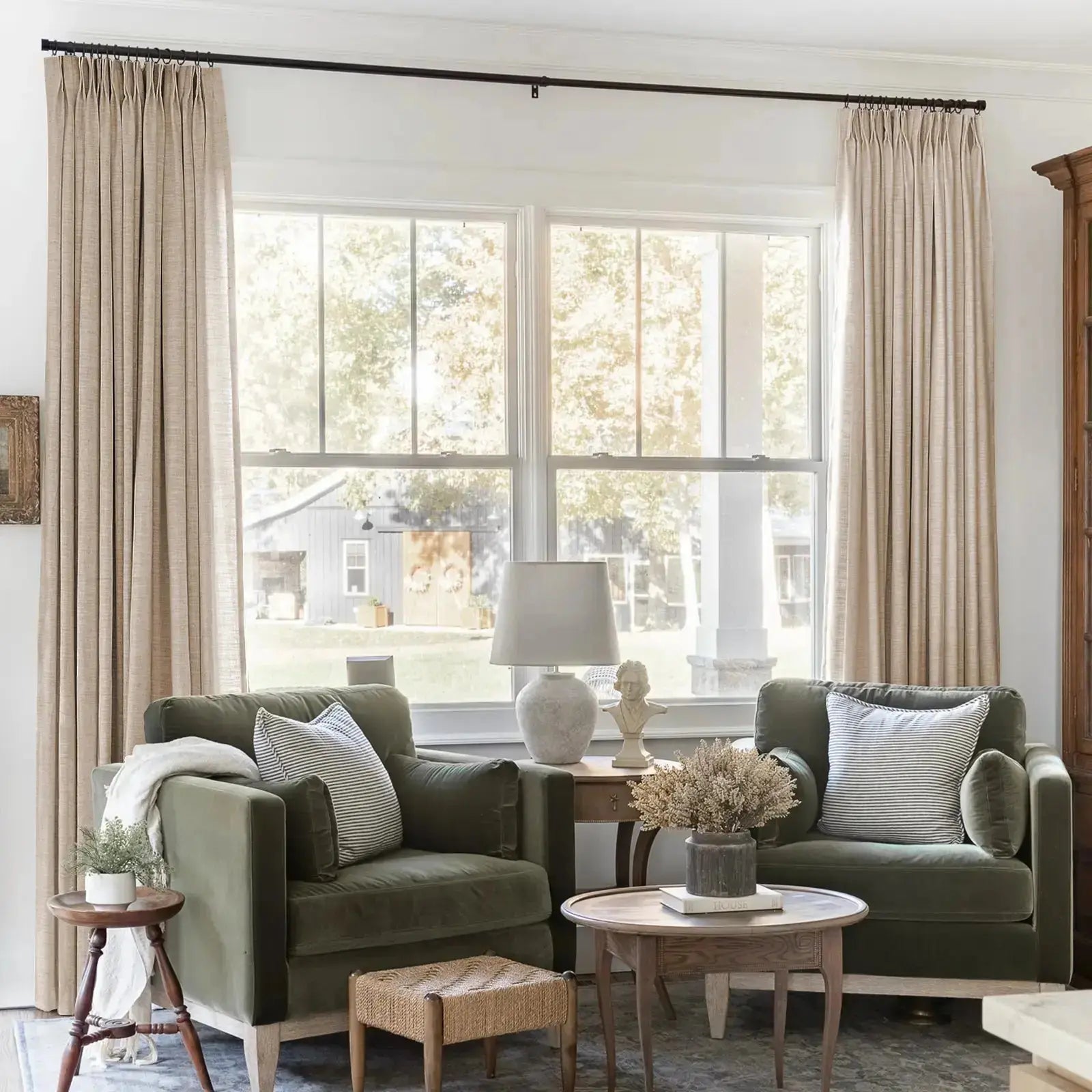 Cozy living room interior with two olive green velvet armchairs and neutral linen curtains framing large windows, offering a view of a rustic barn, featuring a white classical bust and a textured lamp