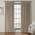 Beige pleated velvet curtains on a black rod frame a large black-grid window, with a grey abstract tree artwork on the left and a cream armchair with a fringed throw blanket on the right