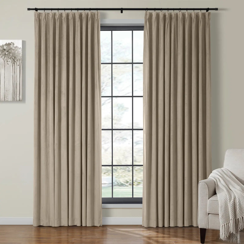 Beige pleated velvet curtains on a black rod frame a large black-grid window, with a grey abstract tree artwork on the left and a cream armchair with a fringed throw blanket on the right