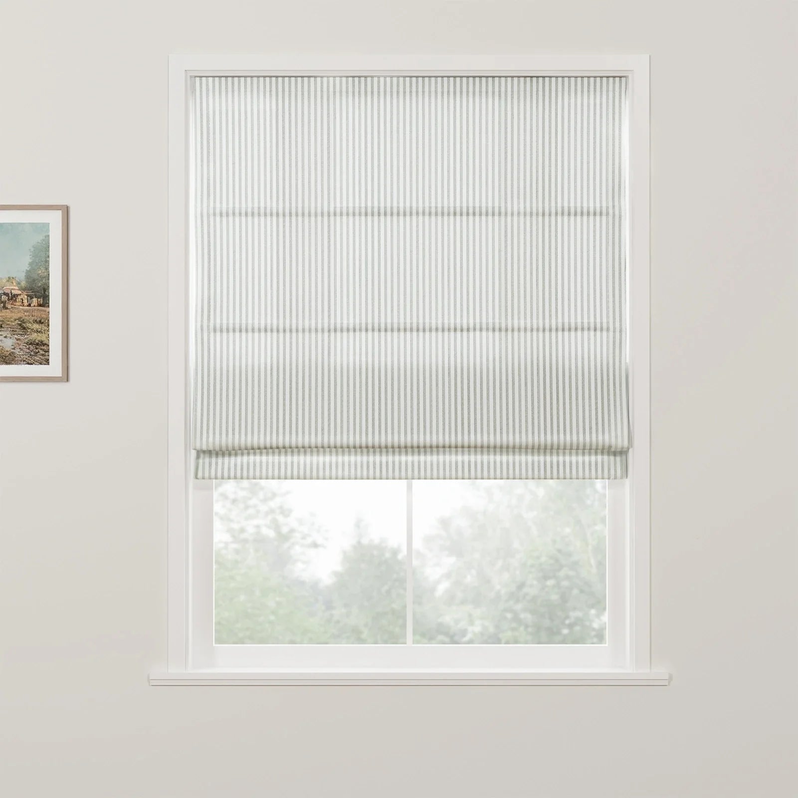 A white window features a grey and white striped roman blind partially raised, revealing blurred green trees outside and a framed landscape artwork on the adjacent wall