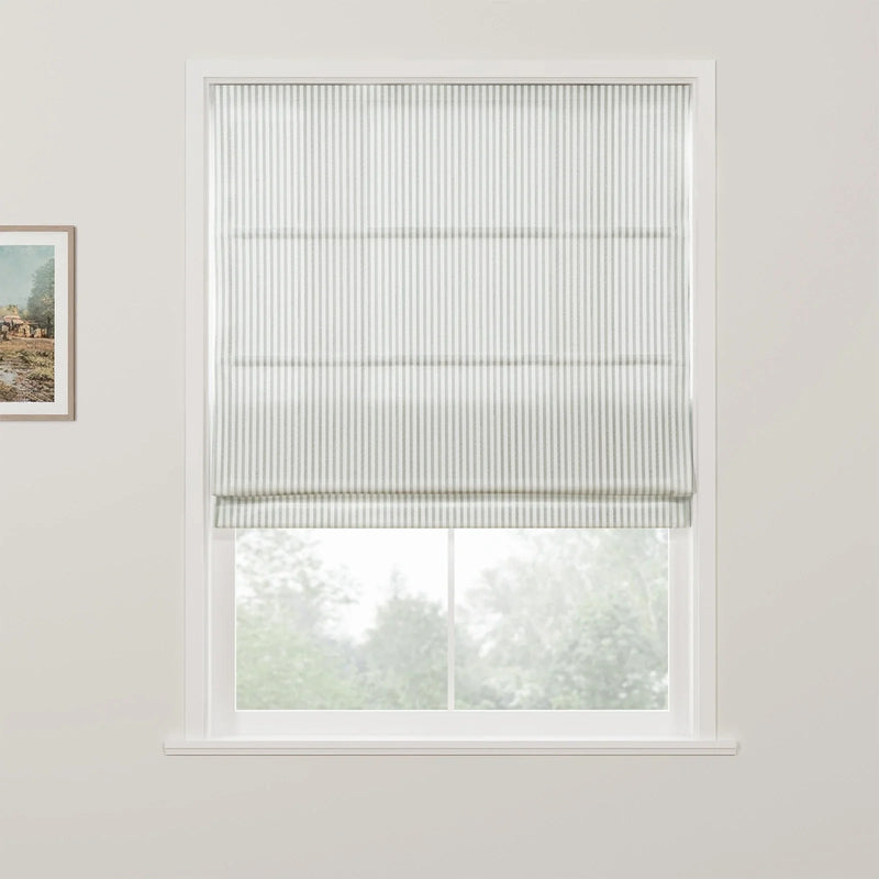 A white window features a grey and white striped roman blind partially raised, revealing blurred green trees outside and a framed landscape artwork on the adjacent wall