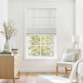 A bright modern room interior features a window with a white pinstripe Roman shade overlooking green trees, a light wood dresser with a pale green vase of dried branches, and a cream upholstered armchair with a soft white throw