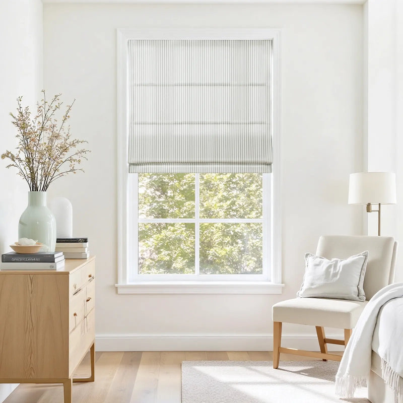 A bright modern room interior features a window with a white pinstripe Roman shade overlooking green trees, a light wood dresser with a pale green vase of dried branches, and a cream upholstered armchair with a soft white throw