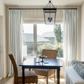 A bright dining nook features a dark wood table and striped curtains, accented by two woven wicker chairs, a bowl of yellow lemons, and a black lantern chandelier overlooking a suburban neighborhood