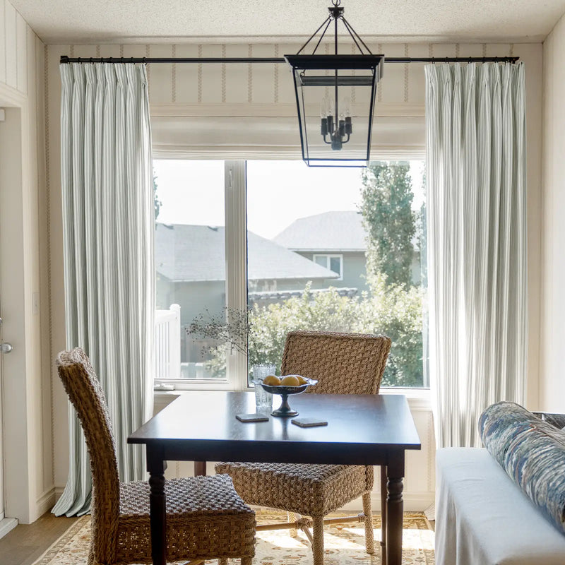 A bright dining nook features a dark wood table and striped curtains, accented by two woven wicker chairs, a bowl of yellow lemons, and a black lantern chandelier overlooking a suburban neighborhood