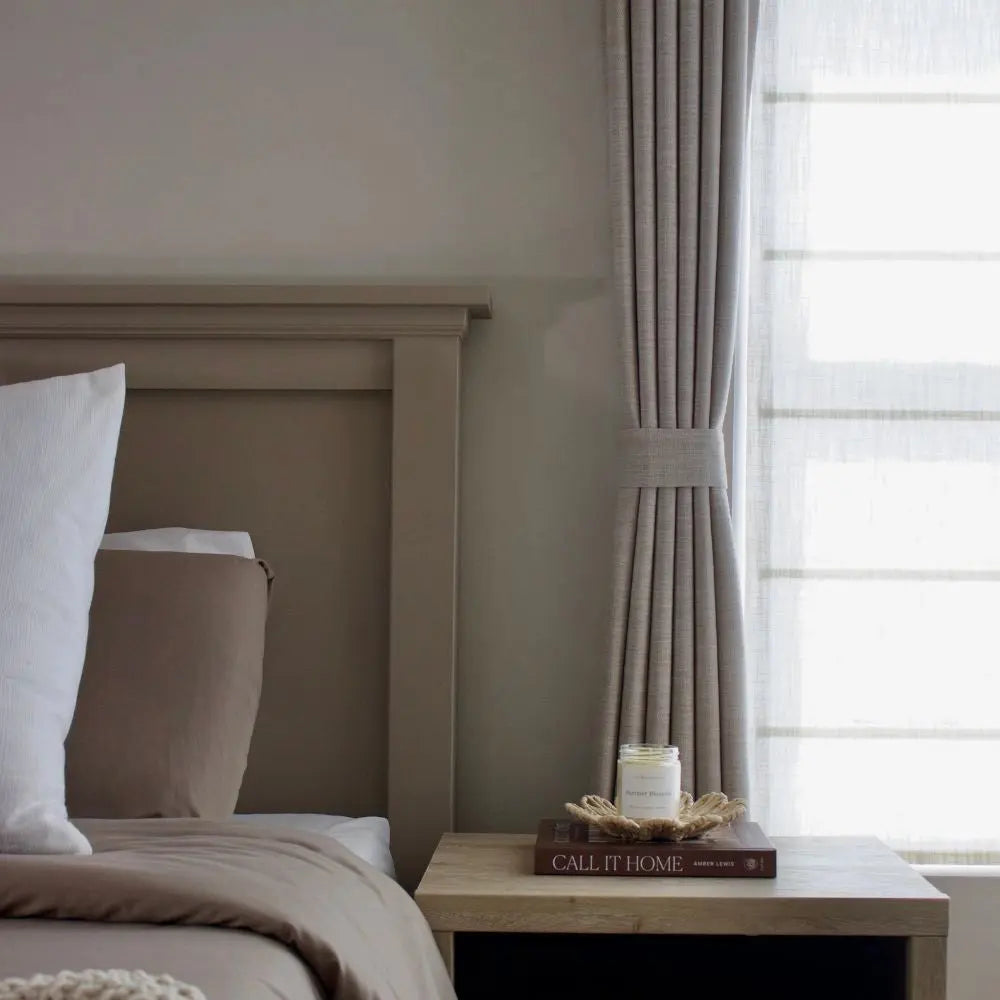 A cozy bedroom interior with a neutral taupe and beige color palette, featuring a wooden headboard, tailored linen curtains tied back, and a light wood nightstand with a "Call It Home" book and a vanilla candle in a woven dish