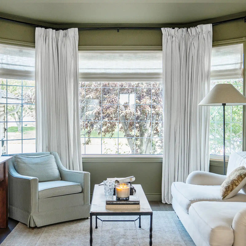 Photography of a cozy living room interior featuring a bay window with white and grey striped curtains, a light blue striped armchair, a white sofa, and a coffee table with a lit candle and books