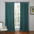Teal blue linen-textured pinch-pleat curtains frame a white paned window revealing sunlit trees, with an ornate cream armchair and a framed lemon botanical print in the room
