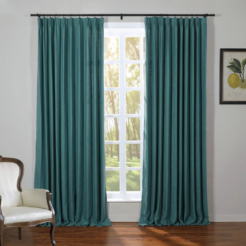 Teal blue linen-textured pinch-pleat curtains frame a white paned window revealing sunlit trees, with an ornate cream armchair and a framed lemon botanical print in the room