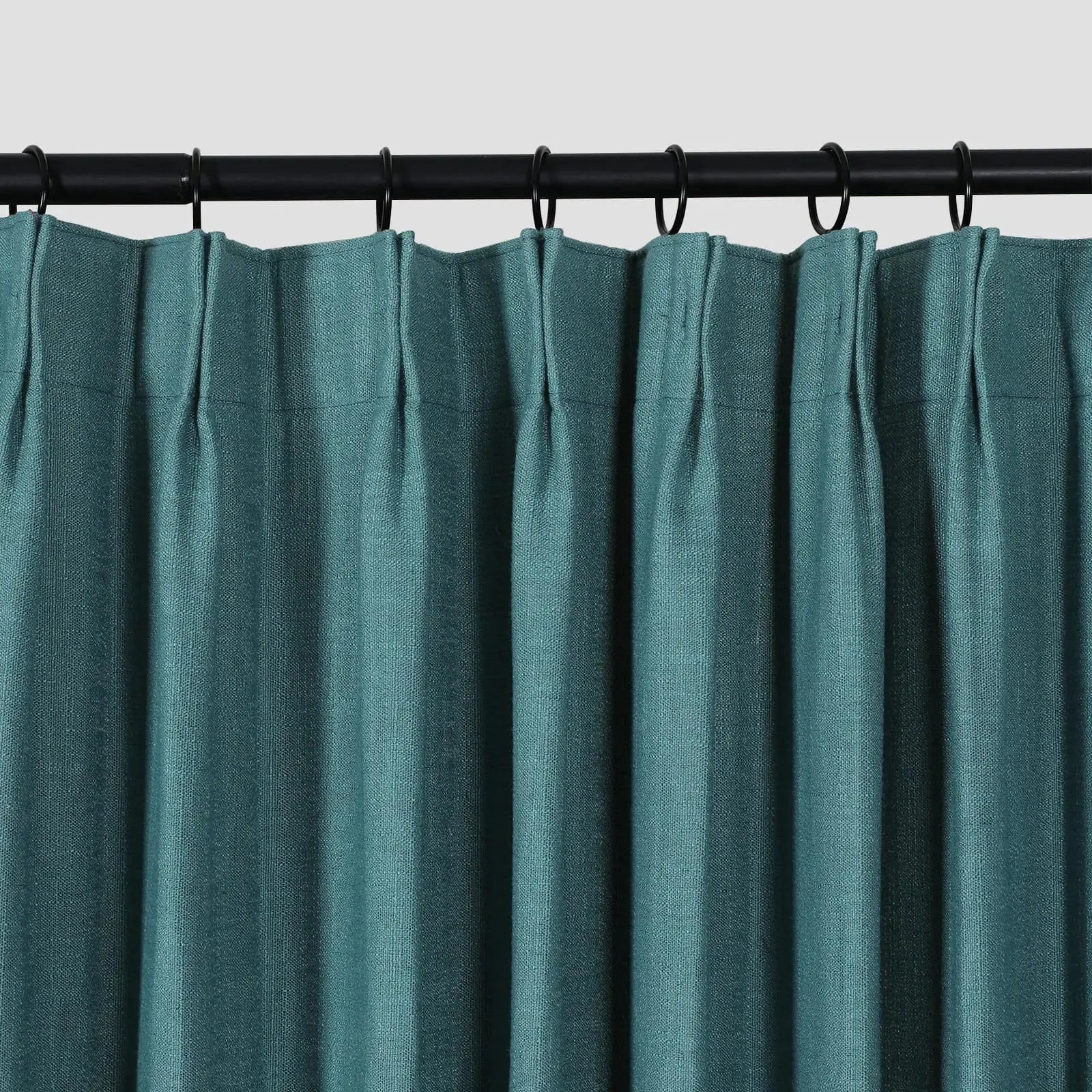 Photograph of a deep teal blue-green textured pleated curtain hung on a black rod with matching black rings