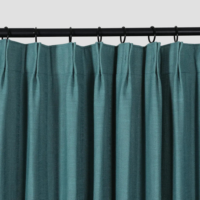 Photograph of a deep teal blue-green textured pleated curtain hung on a black rod with matching black rings