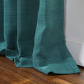 Photograph of a vibrant teal textured curtain with soft pleats pooling elegantly on a polished dark wood floor against a crisp white baseboard
