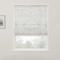 Photograph of a white Roman blind with a subtle grey floral pattern covering the top half of a window in a bright room, revealing blurred green foliage outside, with a small framed picture on the left white wall