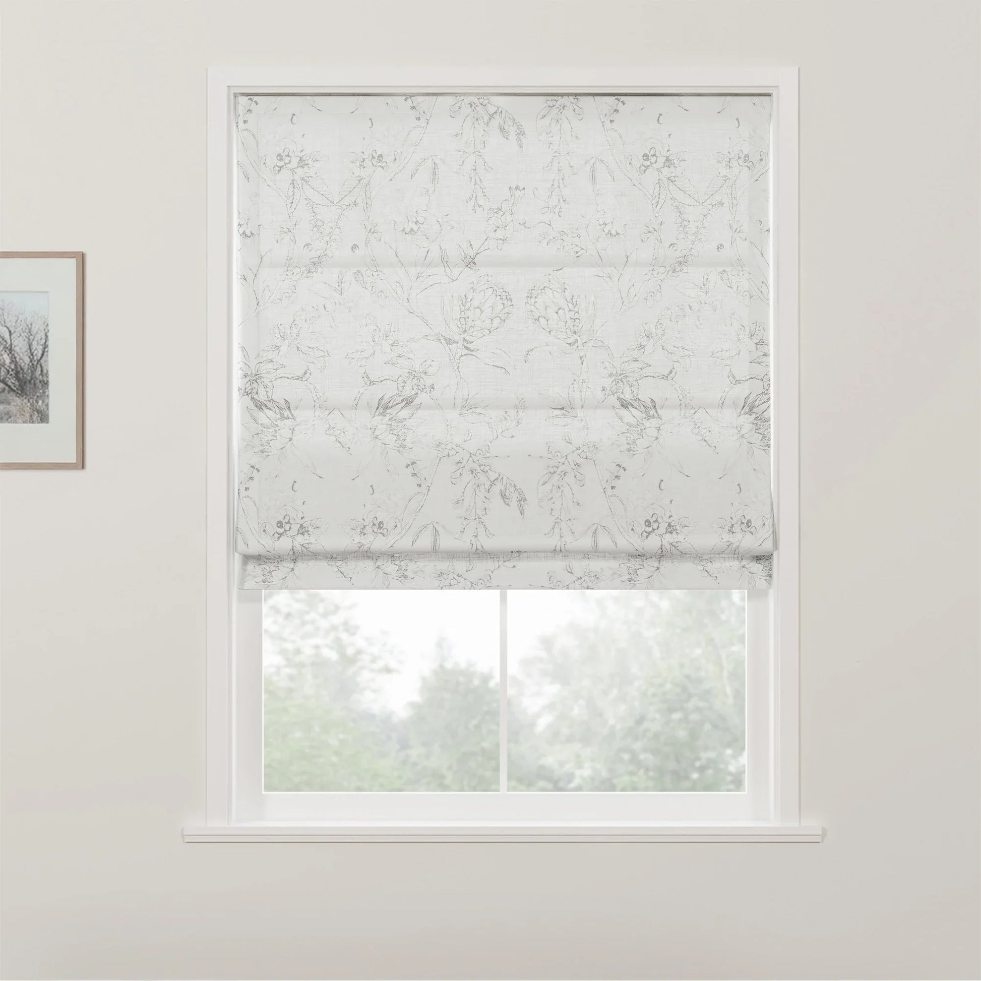 Photograph of a white Roman blind with a subtle grey floral pattern covering the top half of a window in a bright room, revealing blurred green foliage outside, with a small framed picture on the left white wall