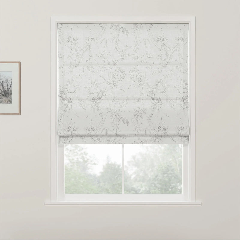 Photograph of a white Roman blind with a subtle grey floral pattern covering the top half of a window in a bright room, revealing blurred green foliage outside, with a small framed picture on the left white wall
