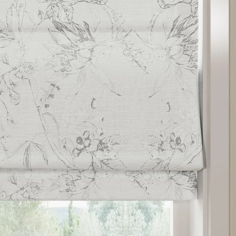 A partially lowered white textured fabric roman blind featuring a monochrome grey botanical sketch pattern of flowers and berries overlooking blurred outdoor greenery