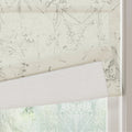 A cream Roman shade with a delicate grey botanical pattern of leaves and flowers partially covers a window revealing its plain white lining and a blurred green outdoor view