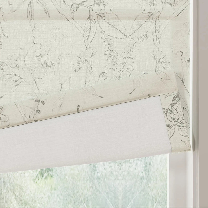 A cream Roman shade with a delicate grey botanical pattern of leaves and flowers partially covers a window revealing its plain white lining and a blurred green outdoor view
