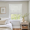 Bright bedroom interior with a window featuring a white floral Roman shade, a light wood bedside table holding a ceramic vase of green branches, and a framed botanical print above white linen bedding