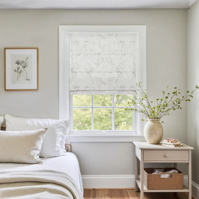 Bright bedroom interior with a window featuring a white floral Roman shade, a light wood bedside table holding a ceramic vase of green branches, and a framed botanical print above white linen bedding