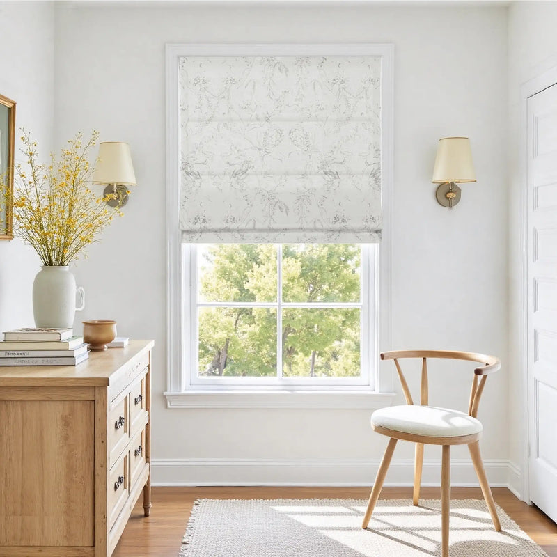 Photograph of a bright, elegant room interior featuring a wooden dresser with a white vase of vibrant yellow flowers, a window with a sheer gray floral patterned Roman shade revealing green trees, and a light wood wishbone-style chair with a cream cushion
