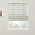 A white window frame showcases a Roman blind with a repeating subtle green leafy pattern partially raised to reveal a blurred natural view of green trees outside, alongside a framed rustic landscape painting on the left wall