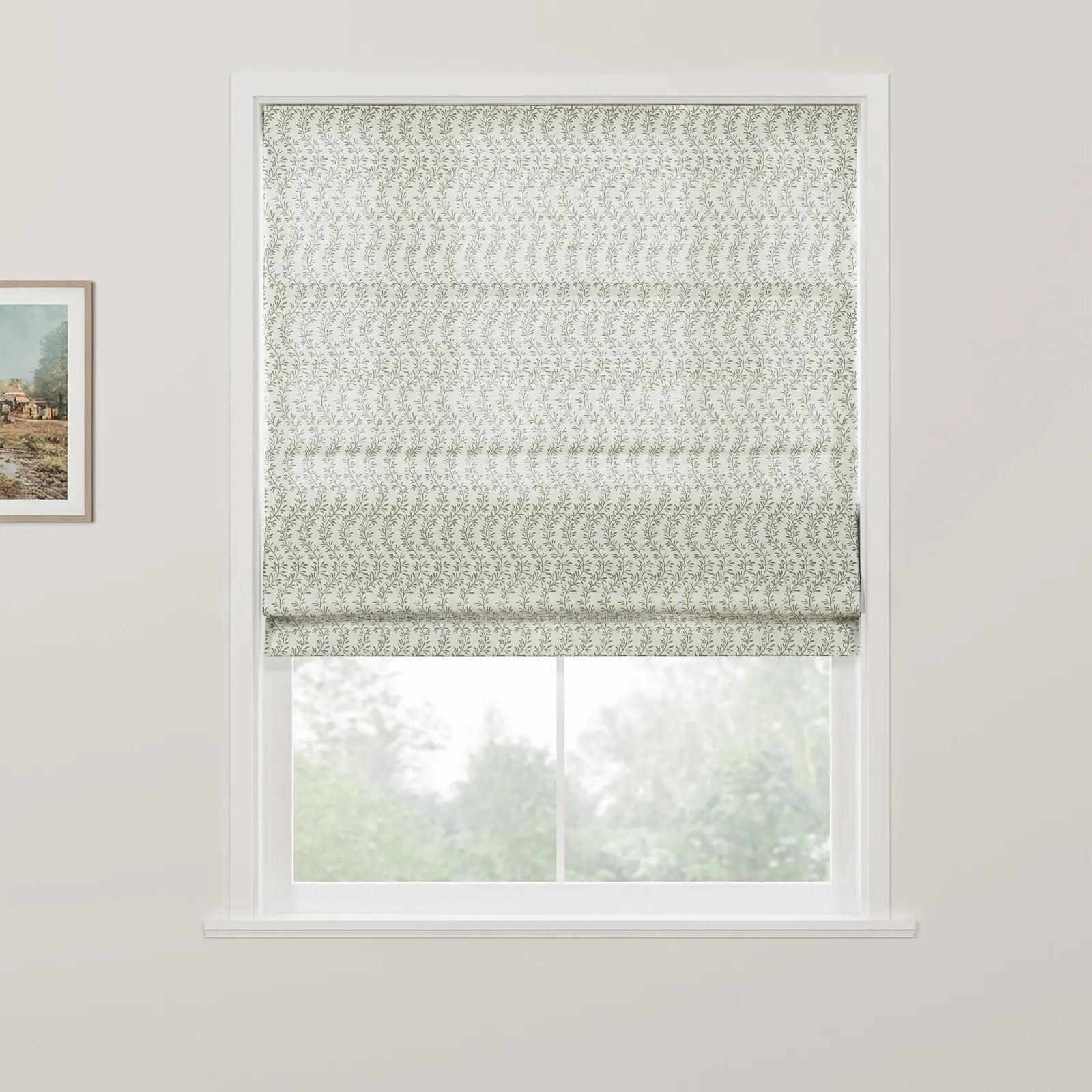 A white window frame showcases a Roman blind with a repeating subtle green leafy pattern partially raised to reveal a blurred natural view of green trees outside, alongside a framed rustic landscape painting on the left wall