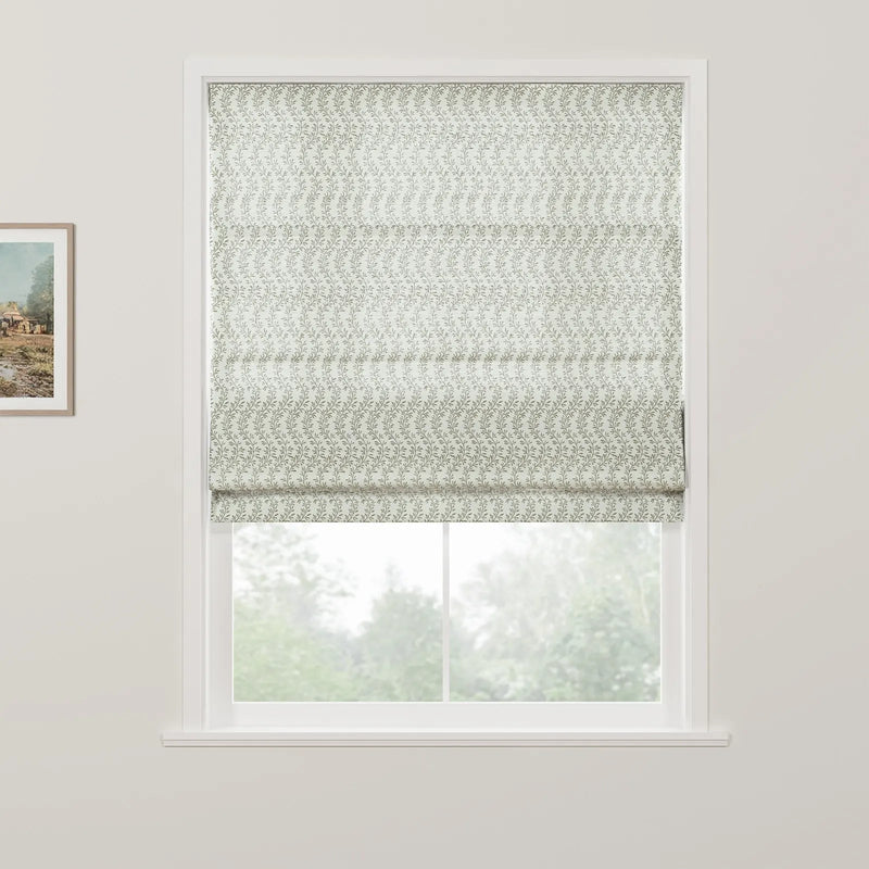 A white window frame showcases a Roman blind with a repeating subtle green leafy pattern partially raised to reveal a blurred natural view of green trees outside, alongside a framed rustic landscape painting on the left wall