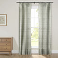 A bright interior features a large window with two pleated drapery panels showcasing a subtle green botanical pattern, a light wood dresser with a framed botanical print sits beside it, offering a view of lush green outdoors