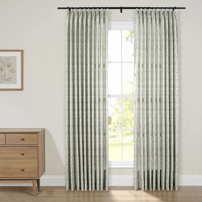 A bright interior features a large window with two pleated drapery panels showcasing a subtle green botanical pattern, a light wood dresser with a framed botanical print sits beside it, offering a view of lush green outdoors