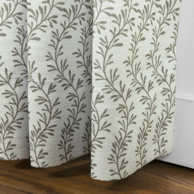 Off-white pleated curtain featuring a repeating brown vine and leaf pattern draped over a dark wooden floor beside a white baseboard
