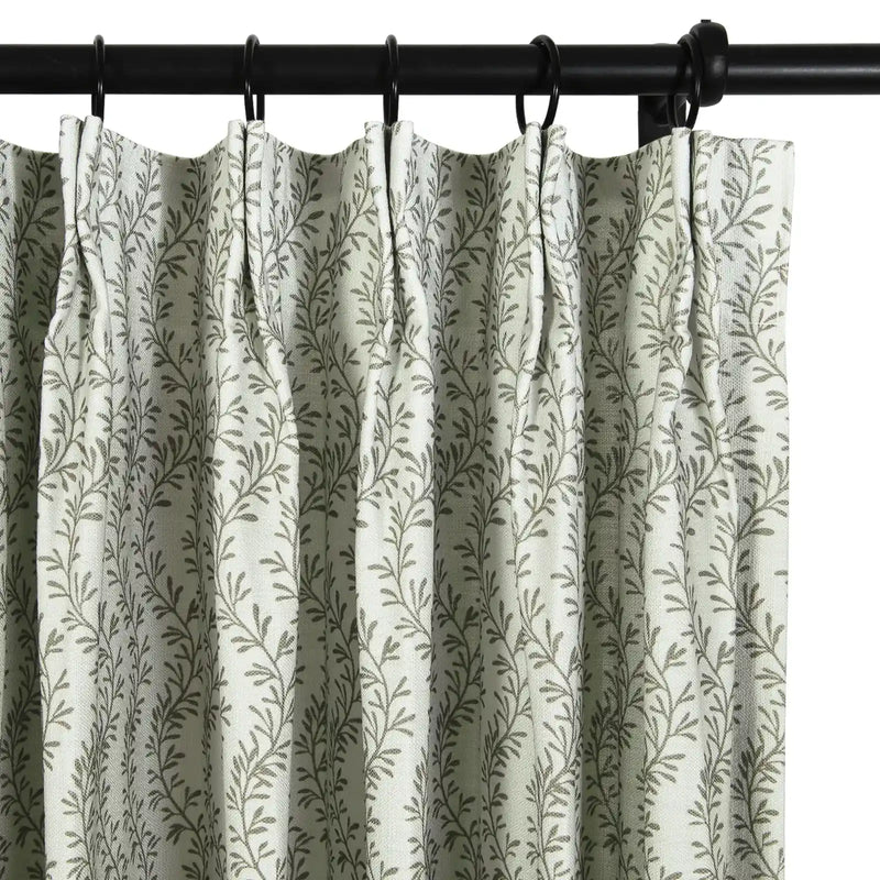 Photograph of a pleated white curtain with a repeating muted green leaf pattern, hung with black rings on a dark curtain rod