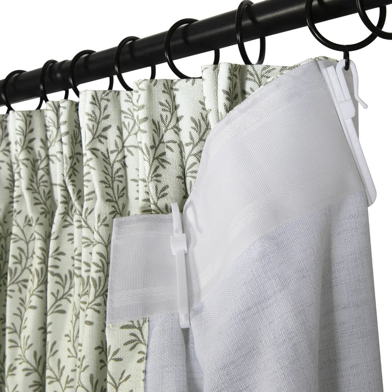 Close-up photograph of a black metal curtain rod with rings holding a pleated light green curtain featuring an olive leaf pattern alongside a white curtain liner secured by plastic hooks