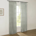 Photograph of a stylish room with white walls, featuring custom pinch pleat curtains in a subtle green botanical pattern on a black rod, beside a framed floral print, with a window view of verdant trees and a hardwood floor