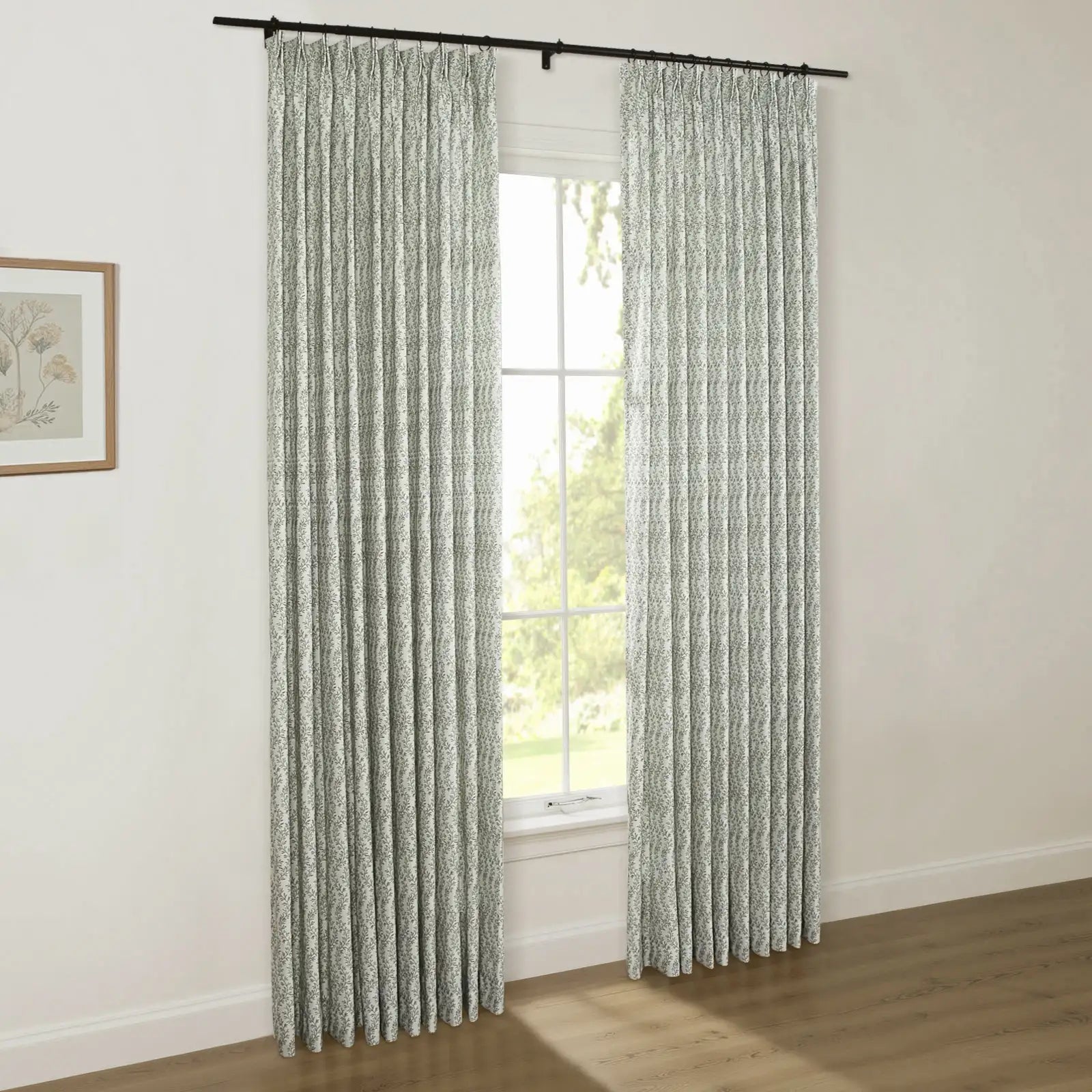 Photograph of a stylish room with white walls, featuring custom pinch pleat curtains in a subtle green botanical pattern on a black rod, beside a framed floral print, with a window view of verdant trees and a hardwood floor