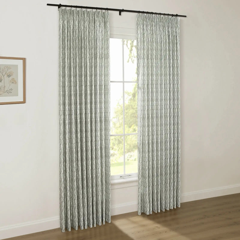 Photograph of a stylish room with white walls, featuring custom pinch pleat curtains in a subtle green botanical pattern on a black rod, beside a framed floral print, with a window view of verdant trees and a hardwood floor