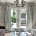 Photography of a bright living room interior featuring white French doors framed by long pleated green leaf pattern curtains, a modern circular chandelier with six white shades, and a glass coffee table with blue trim displaying fabric swatches, overlooking an outdoor patio with concrete elements and lush green trees